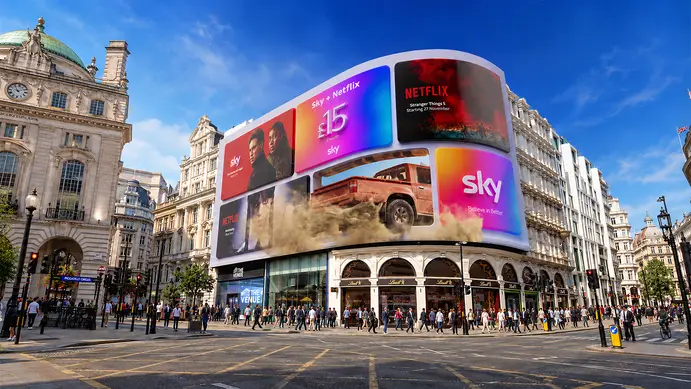 The curved Piccadilly Lights screen above a busy Piccadilly Circus on a sunny day, showing Sky and Netflix adverts